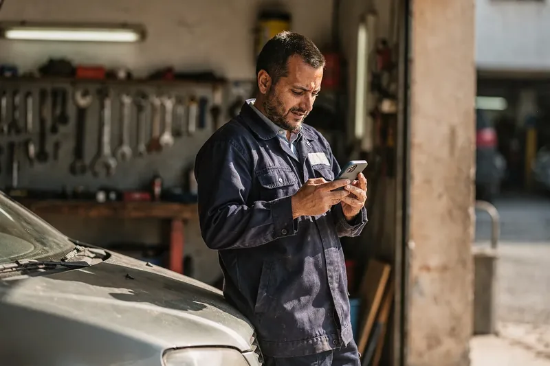 Mecánico apoyado en un coche leyendo el móvil en su taller durante un descanso